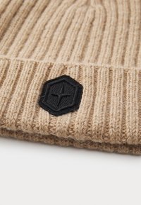 Căciulă tricotată cu rimă bej, cu un patch hexagonal negru care prezintă un design embosat cu o stea. Textură moale și ajustare flexibilă.