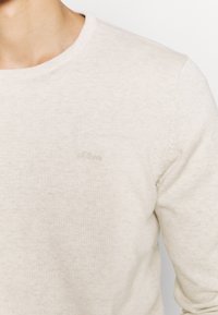 Hellbeiger Pullover aus strukturiertem Strickstoff mit rundem Ausschnitt. Verfügt über ein dezentes, gesticktes Logo oben links.