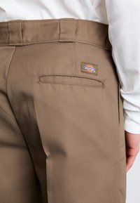 Hnědé bavlněné kalhoty s hladkou texturou, s jednou zadní kapsou a malou nášivkou loga Dickies na pásku.