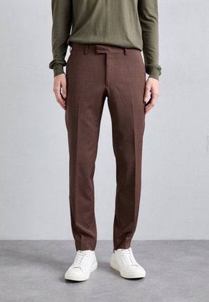 Pantalones cortos marrones de corte ajustado, hechos de una tela ligera, con un frente plano y detalles de pliegue sutiles, combinados con zapatillas blancas.
