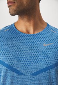 Sinine spordipoolsärk, mis on valmistatud tekstuuriga kangast, geomeetrilise mustriga, ümmarguse kaelusega ja sellel on rinnal väike hall Nike logo.