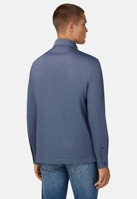 Langermige top van donkerblauwe stof met een kraag, voorzien van knoopsluitingen bij de manchetten en een soepele textuur, gecombineerd met lichtblauwe jeans.