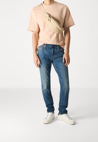 Ljusrosa skjorta med korta ärmar, blå jeans och vita sneakers. En beige axelväska bärs tvärs över bröstet. Avslappnad, ledig passform.