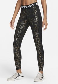 Leggings negras con patrón de hojas doradas, cinturilla elástica etiquetada "NIKE PRO", diseño ajustado, combinados con zapatillas deportivas blancas.
