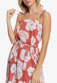 Mujer ajustando la correa de un vestido sin mangas de color óxido con un patrón de grandes flores y hojas tropicales blancas, atado en la cintura.