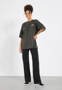 Donkergroene oversized t-shirt met korte mouwen en geborduurde details, gecombineerd met zwarte wijde broek en marineblauwe sneakers.