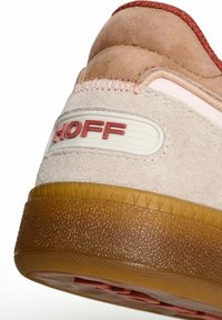Zapatillas con una parte superior de ante beige y acentos en rosa, una suela de goma marrón texturizada y detalle de marca en el talón en rojo.