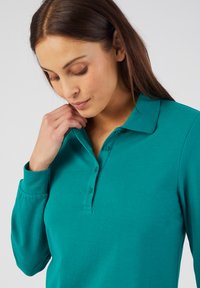 Camisa de polo de manga larga turquesa con cuello, tapeta de tres botones y textura de tela suave. Vista en primer plano, detalles visibles mínimos.