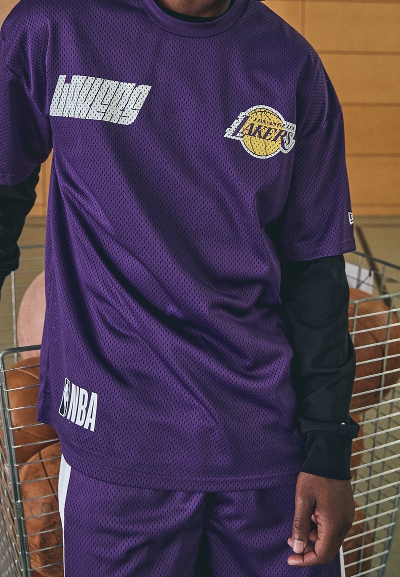 Osoba ubrana w fioletową siateczkową koszulkę NBA Los Angeles Lakers i pasujące spodenki, stojąca wewnątrz obiektu obok metalowego kosza na przechowywanie.