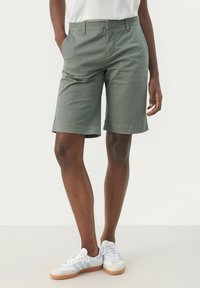 Shorts en coton vert à coupe droite, avec poches latérales et ourlet replié. Portés avec des baskets blanches et grises.