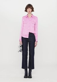 Pink satin shirt with two chest pockets, paired with dark denim jeans. Model holds a small silver metallic bag, wearing black boots.