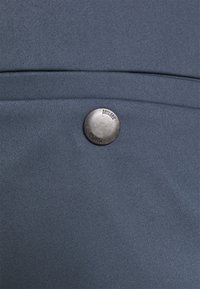 Mörkgrått tyg med en slät textur, som har en rund metallknapp graverad med "DRYKORN" nära övre vänstra hörnet.