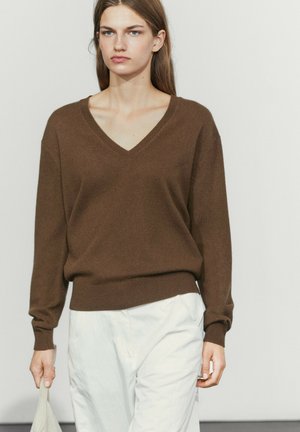 Femme portant un pull marron col en V et un pantalon blanc tenant un sac beige sur un fond uni.
