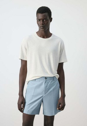 Homme portant une chemise blanche à manches courtes unie et un short bleu clair sur mesure, debout devant un fond clair uni.