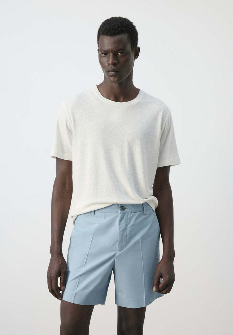 Homme portant une chemise blanche à manches courtes unie et un short bleu clair sur mesure, debout devant un fond clair uni.