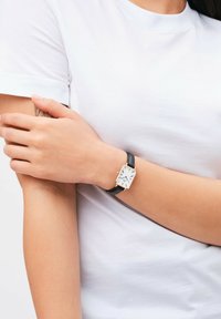 Black leather strap watch with a rectangular silver case. White dial features black hour markers and hands. Worn on a bare wrist.