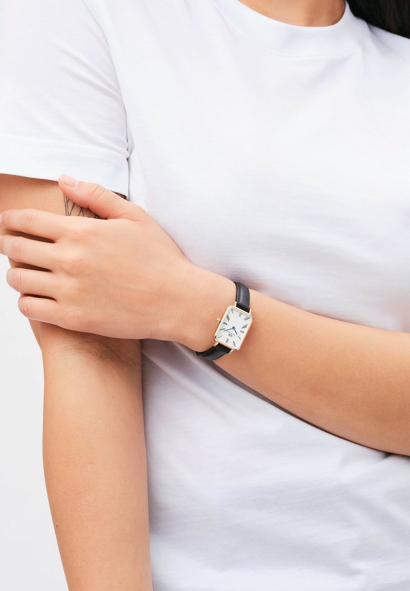 Black leather strap watch with a rectangular silver case. White dial features black hour markers and hands. Worn on a bare wrist.