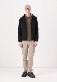 Sort zip-up jakke, beige polo, lyse beige bukser og mørkegrå sneakers, båret af en person, der står foran en neutral baggrund.