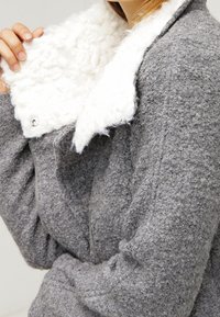 Grå texturerad jacka med vit konstpälskrage, med dragkedja framtill och sidodetaljer för en väldefinierad passform.