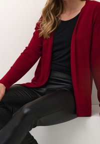 Rode cardigan met een getextureerde breiwijze, open voorkant, gedragen over een zwart topje en zwarte broek met leerachtige uitstraling, met een gladde stof en een aansluitende pasvorm.