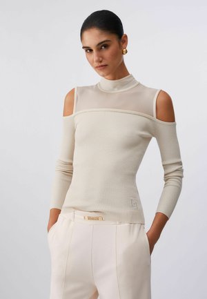 Donna che indossa un maglione crema a coste con spalle scoperte, collo alto trasparente, orecchini dorati e pantaloni crema chiaro con dettaglio cintura a catena.