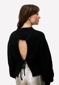 Zwarte trui van pluizig materiaal, met een diepe uitsnijding aan de achterkant en een striksluiting, brede mouwen en een ontspannen pasvorm. Zilveren broek zichtbaar.