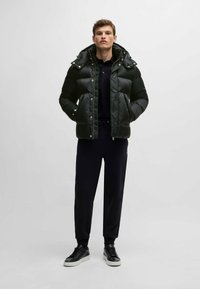 Svart puffjacka med texturerad design, utrustad med avtagbar huva, tryckknappar och sidofickor, kombinerad med svarta byxor och sneakers.