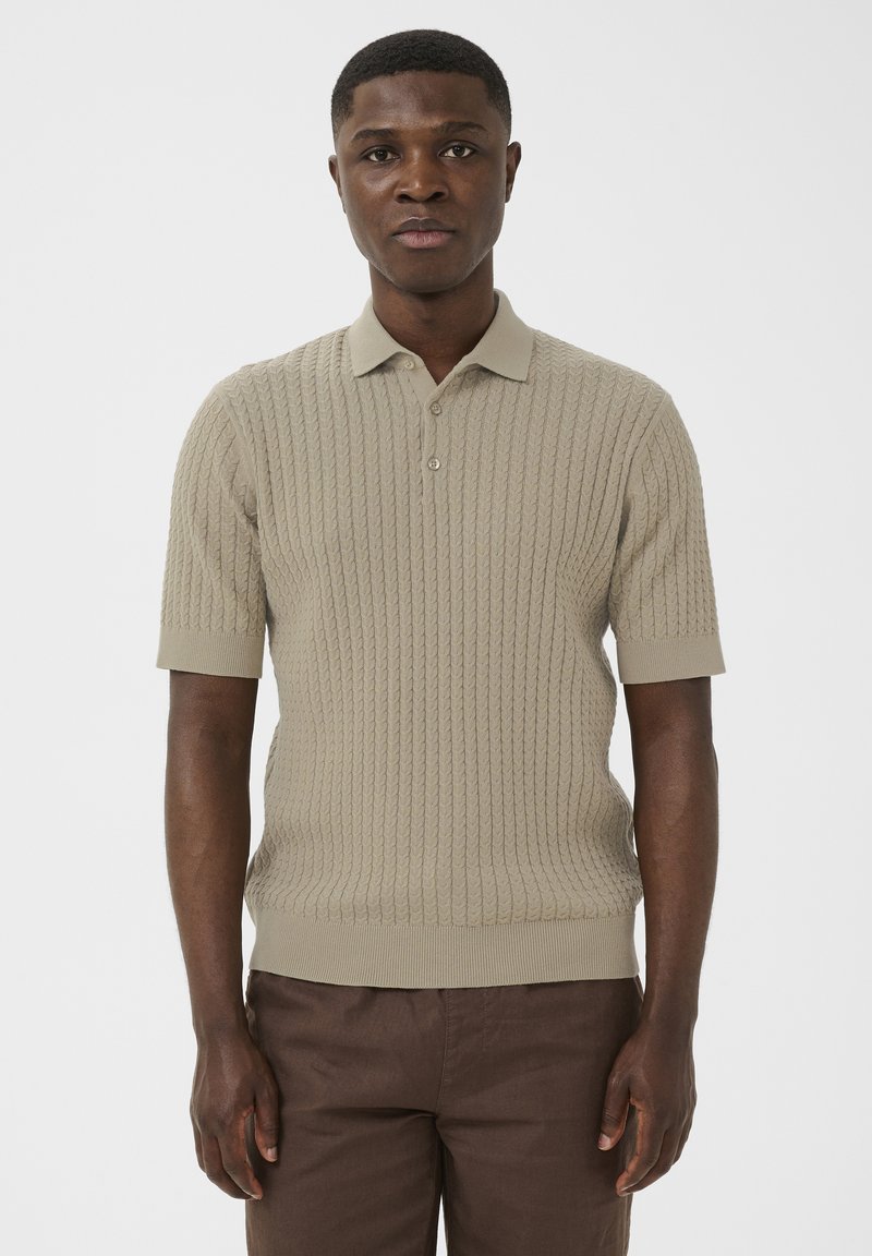 Homme debout portant un polo en maille torsadée beige à manches courtes et un pantalon marron, sur un fond blanc uni.