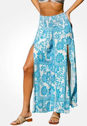Vrouw draagt wit-blauwe wijde broek met bloemenpatroon, hoge splits aan beide zijden en gevlochten sandalen.