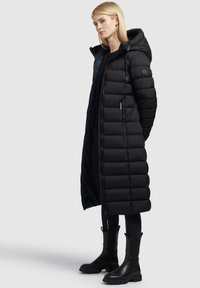 Fekete puffernadrág hosszú sziluettel, steppelt dizájnnal és kapucnival; cipzáras zsebekkel és sima belső béléssel rendelkezik. Fekete bokacsizma tartozik hozzá.