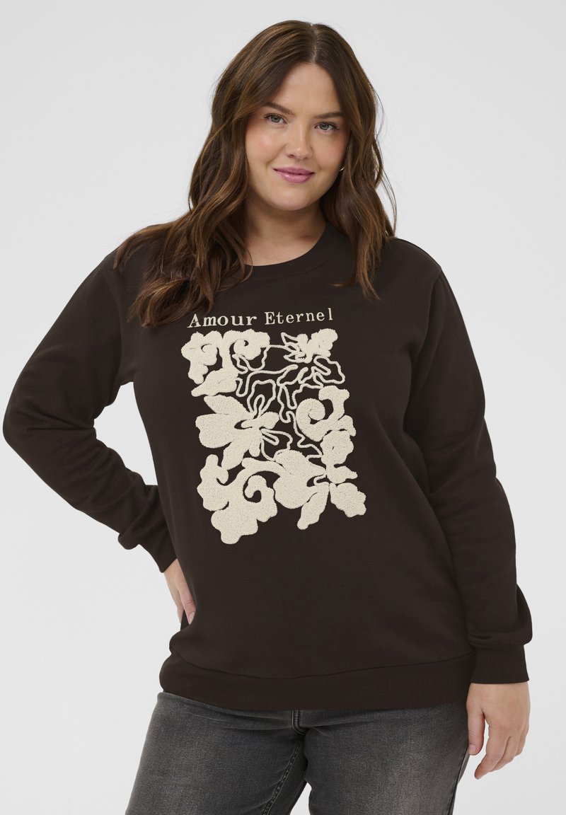 Femme aux longs cheveux bruns portant un sweat-shirt noir avec un motif floral beige et le texte « Amour Eternel », posant devant un fond uni.