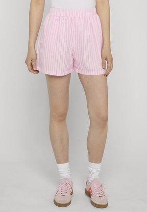 Person trägt hellrosa-weiß gestreifte Shorts, weiße Socken und pinke Sneakers, steht vor einem einfarbigen grauen Hintergrund.