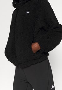 Giacca in fleece nera con una superficie texturizzata, dotata di un colletto alto e un patch con logo. Indossata con leggings neri.