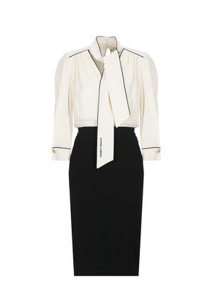 Blusa bianca con colletto annodato, bordi neri e maniche a sbuffo; abbinata a una gonna a matita nera a vita alta con dettagli dorati.