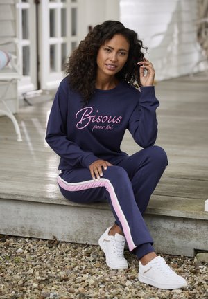 Marineblauw sweatshirt met roze geborduurde tekst en bijpassende sweatpants met roze en witte zijkanten, gecombineerd met witte sneakers.