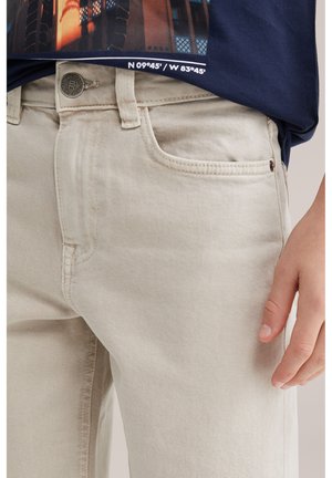 Persona vistiendo jeans beige claro y una camisa azul marino con un paisaje urbano y coordenadas impresas en el dobladillo.