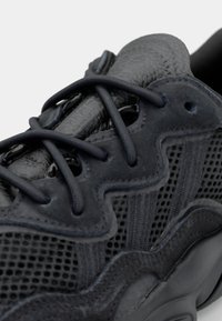 Zapatilla gris oscura con una parte superior texturizada que incluye paneles de malla, intrincados detalles ondulados y ojales y cordones negros brillantes. Diseño ligero.