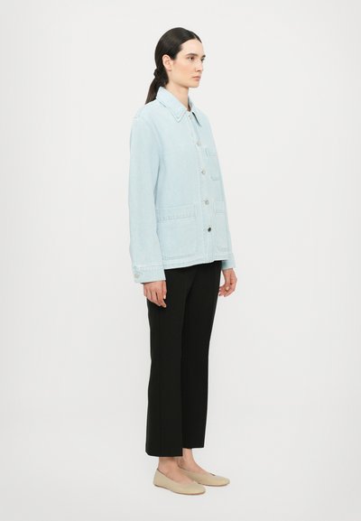 Light blue denim jacket with a collar, front buttons, and two large pockets, paired with black trousers and beige slip-on shoes.