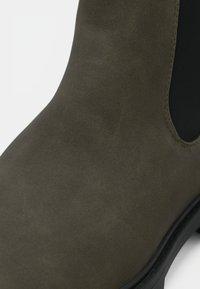 Olivgrüner Knöchelstiefel aus glattem Wildleder, ausgestattet mit elastischen Seitenpaneelen, runder Zehenpartie und einer schwarzen Gummisohle.