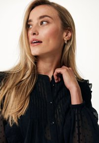 Blusa nera a maniche lunghe con texture a righe, polsini arricciati e chiusura con bottoni. Il modello ha capelli biondi lisci e orecchini a cerchio dorati.