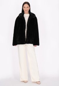 Sort faux fur jakke med sjalkrave, åben foran og løs pasform, kombineret med cremede brede bukser og lyse sko.