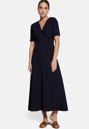 Femme portant une robe portefeuille noire à manches courtes avec une ceinture, des sandales marron à enfiler et des bijoux minimalistes sur un fond uni.