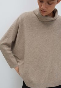 Frau in einem locker sitzenden, beigen Rollkragenpullover mit langen Ärmeln, die nach unten schaut, mit einer Hand auf der Hüfte gegen einen hellen Hintergrund.