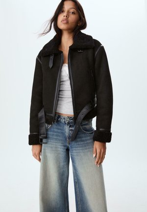 Donna che indossa una giacca in shearling nera con ampi revers e polsini sopra una canotta bianca, abbinata a jeans svasati con vita alta e effetto sbiadito.