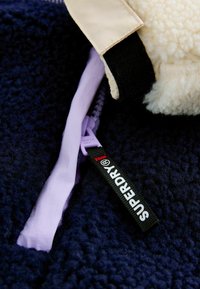 Mornarická flis jakna s mekom, teksturiranom površinom. Ima kremast kragnu, ljubićasti patentni zatvarač i crnu oznaku s bijelim tekstom koji glasi "SUPERDRY®."