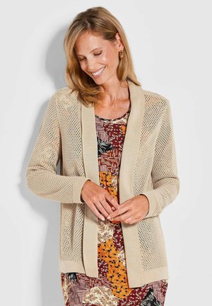 Gilet en maille beige avec un design à mailles ouvertes, des poignets et un col unis. Porté sur une robe à motifs multicolores présentant des motifs floraux et géométriques.
