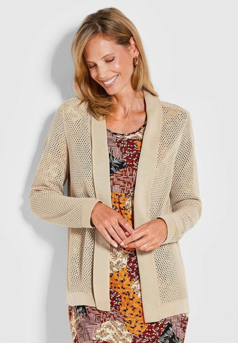 Gilet en maille beige avec un design à mailles ouvertes, des poignets et un col unis. Porté sur une robe à motifs multicolores présentant des motifs floraux et géométriques.