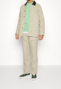 Beige canvas jacket with black collar, green sweatshirt layered underneath, beige pants, and black sneakers featuring a green graphic design.