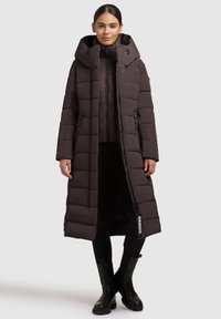 Cappotto imbottito lungo di colore marrone scuro con un ampio cappuccio, design trapuntato, zip anteriore, due tasche laterali e dettaglio del logo sul lato sinistro.