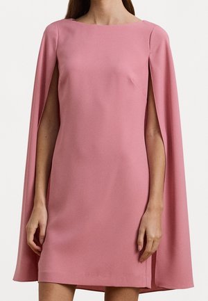 Woman wearing pink dress with long cape sleeves and boat neckline, standing against plain light background.